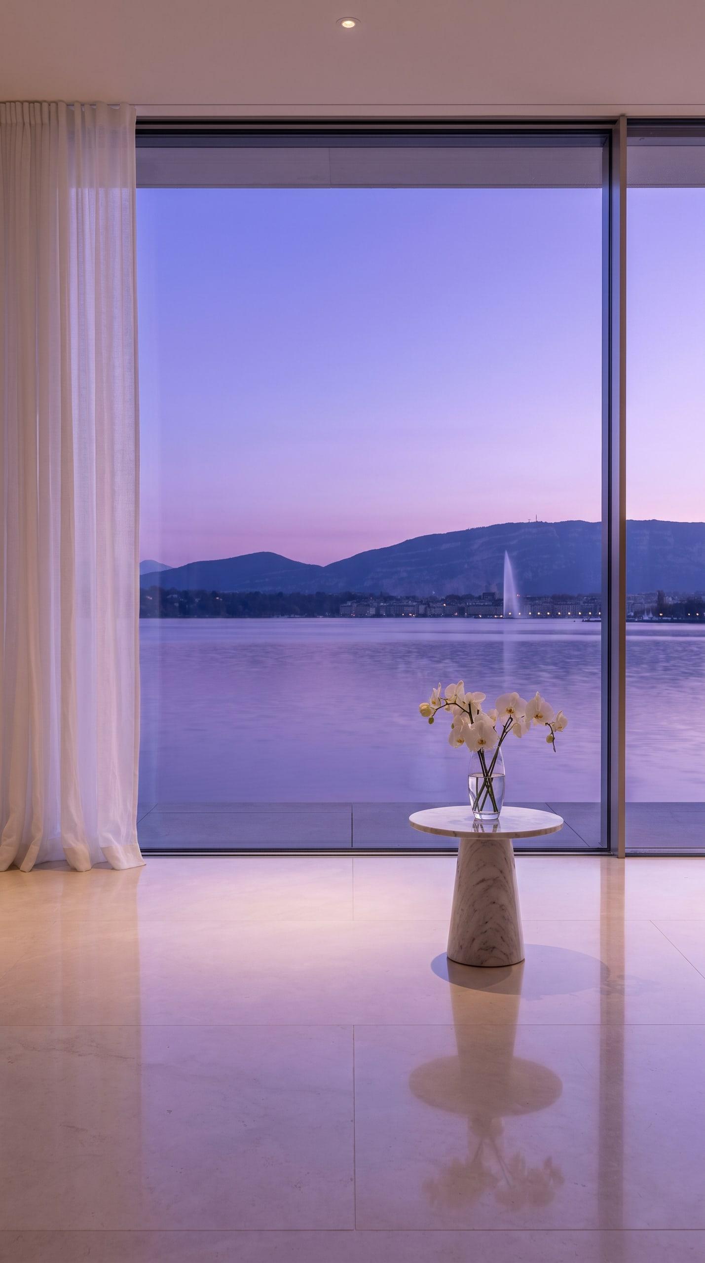 Baie vitrée d'une villa ouvrant sur le lac Léman et le Jet d'eau au crépuscule